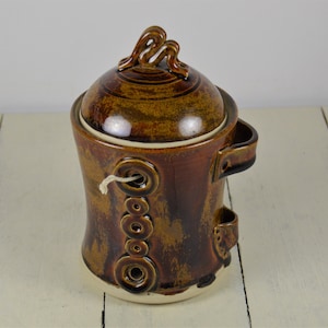 May include: A brown ceramic jar with a lid and a handle. The jar has a decorative design of circles and a string running through the holes. The lid has a decorative design of a swirl.