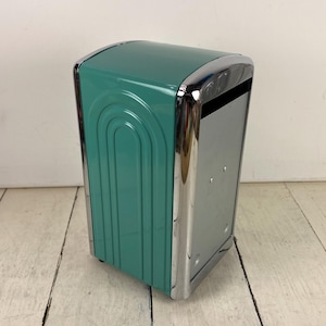 May include: A teal metal napkin dispenser featuring a shiny chrome frame and a curved, embossed arch design on the side panels. The dispenser has a vertical orientation with a rectangular opening at the top for dispensing napkins from the front.