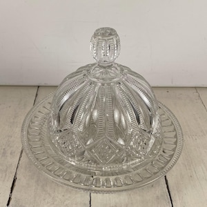 May include: A clear, vintage glass butter dish with a domed lid and a round base. The lid features a decorative knob and a ribbed design, while the base has a patterned edge. The dish is made of clear glass.
