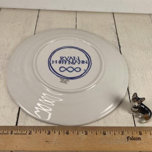 K&ouml;nnte beinhalten: Ein wei&szlig;er Keramikteller mit blauem Logo "RYAN HOFFMANN" und Unendlichkeitszeichen. Eine Miniatur-Corgi-Figur und ein Holzlineal sind ebenfalls vorhanden. Der Teller hat einen Durchmesser von etwa 15 cm.
