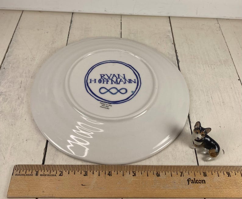 K&ouml;nnte beinhalten: Ein wei&szlig;er Keramikteller mit blauem Logo "RYAN HOFFMANN" und Unendlichkeitszeichen. Eine Miniatur-Corgi-Figur und ein Holzlineal sind ebenfalls vorhanden. Der Teller hat einen Durchmesser von etwa 15 cm.