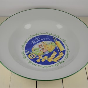 May include: A white ceramic pasta bowl with a green rim and a blue and green illustration of pasta, a person, and the text "La Pasta" in the center. The text around the illustration reads "Rottelle - Lasagne - Spaghetti - Tagliatelle -  Italiani - Orecchiette -  Fusilli - Ravioli -  Tortellini -  Gnocchi".
