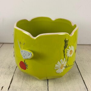 Może przedstawiać: Chartreuse ceramiczna donica z falistym brzegiem. Donica jest ozdobiona motylem, czerwoną truskawką i białymi stokrotkami. Donica to element dekoracyjny do domu.
