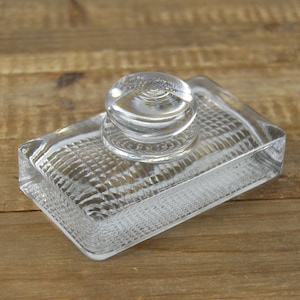 May include: A clear glass inkwell with a textured surface and a round glass stopper.