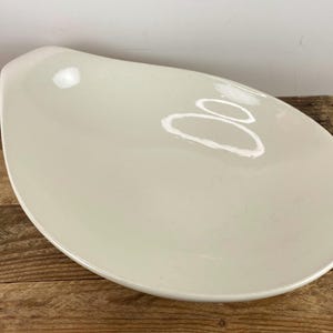 May include: A large, white ceramic serving dish with a unique, wave-like shape. The dish is smooth and glossy, with a slightly off-white color.