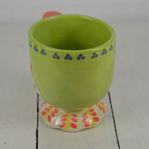 May include: A green ceramic mug with a red handle and a colorful floral design on the bottom. The mug has a white rim and blue dots around the top.