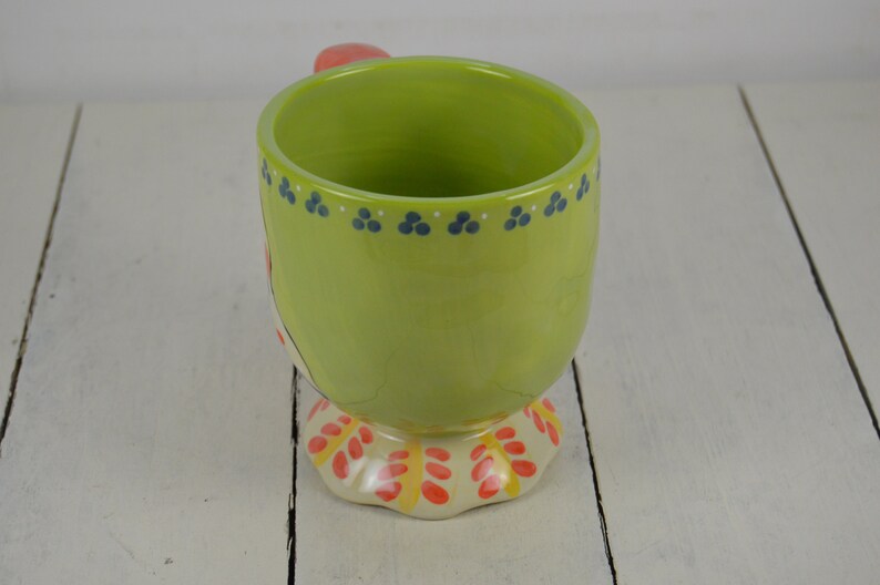 May include: A green ceramic mug with a red handle and a colorful floral design on the bottom. The mug has a white rim and blue dots around the top.