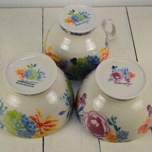 May include: Three white ceramic bowls with floral designs. The bowls are decorated with colourful flowers and leaves. The bottom of each bowl has the word "Anthropologie" printed on it.