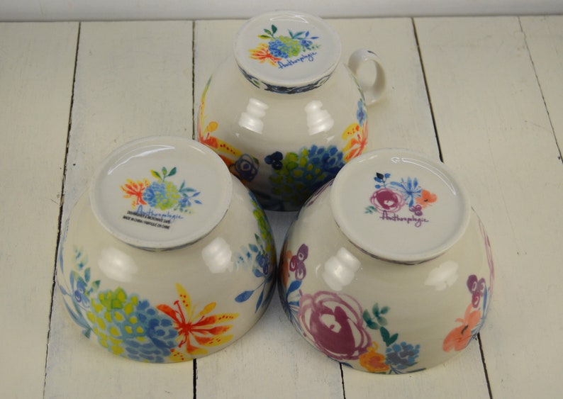 May include: Three white ceramic bowls with floral designs. The bowls are decorated with colourful flowers and leaves. The bottom of each bowl has the word "Anthropologie" printed on it.