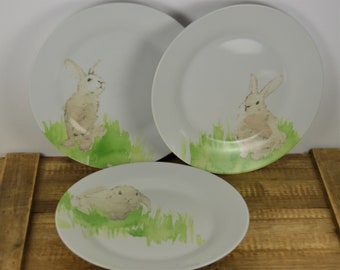 May include: Three white ceramic dinner plates with a watercolor design of bunnies in green grass.