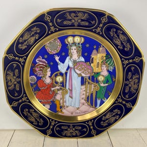 May include: Decorative octagonal plate with a dark blue border and gold trim. The plate features a central illustration of three figures in period clothing, holding candles and a sign that reads "1979". The plate has a gold rim and ornate gold detailing.