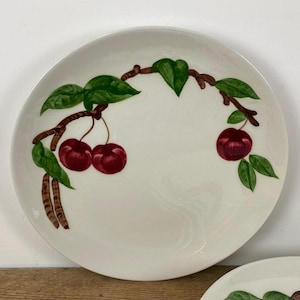 May include: A white ceramic plate with a hand-painted design of cherries on a branch. The cherries are a deep red, with green leaves and brown branches. The plate has a simple, elegant design, perfect for serving food.