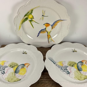 May include: Three white ceramic plates with scalloped edges. Each plate features a hand-painted bird design in vibrant colors, including yellow, blue, and orange. One plate has a dragonfly, and the other two have bees.