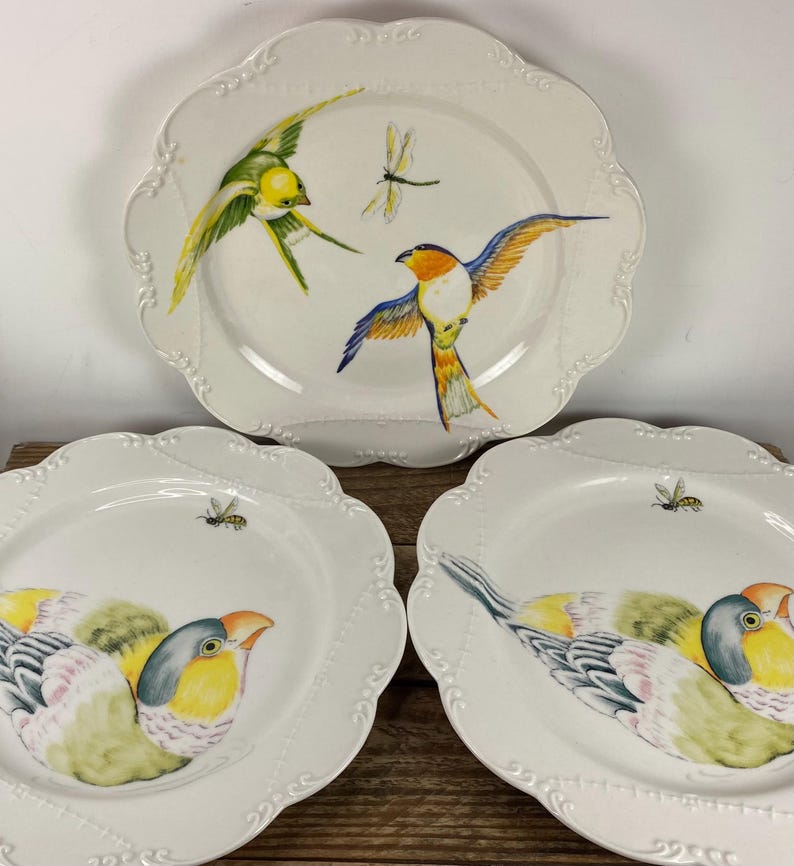May include: Three white ceramic plates with scalloped edges. Each plate features a hand-painted bird design in vibrant colors, including yellow, blue, and orange. One plate has a dragonfly, and the other two have bees.
