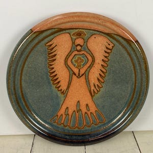 May include: Round ceramic plate with a hand-painted angel design. The angel is in shades of orange and is centered on a teal background. The rim is a darker blue-green with an orange top edge. The angel holds a cross.