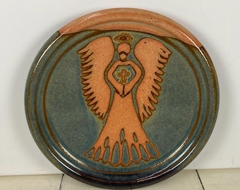 May include: Round ceramic plate with a hand-painted angel design. The angel is in shades of orange and is centered on a teal background. The rim is a darker blue-green with an orange top edge. The angel holds a cross.