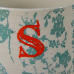 May include: White ceramic mug with a teal floral pattern and a red letter "S" on the front.