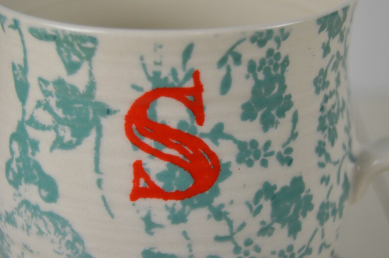 May include: White ceramic mug with a teal floral pattern and a red letter "S" on the front.