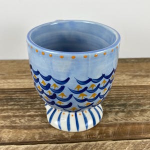 Op de afbeelding: Een keramische mok met een lichtblauwe achtergrond en een golfpatroon in donkerblauw en geel. De rand heeft oranje stippen en de basis is wit met blauwe strepen. De mok staat op een houten oppervlak.