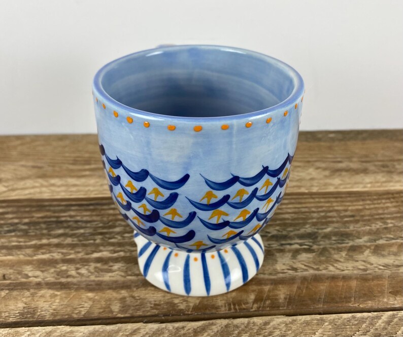 Op de afbeelding: Een keramische mok met een lichtblauwe achtergrond en een golfpatroon in donkerblauw en geel. De rand heeft oranje stippen en de basis is wit met blauwe strepen. De mok staat op een houten oppervlak.