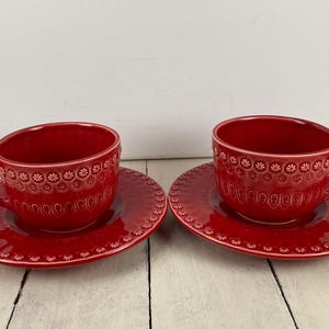 May include: Two matching red ceramic teacups and saucers. The cups and saucers have a floral embossed design around the rim. The cups have a handle and sit on matching round saucers. The set is on a white wooden surface.