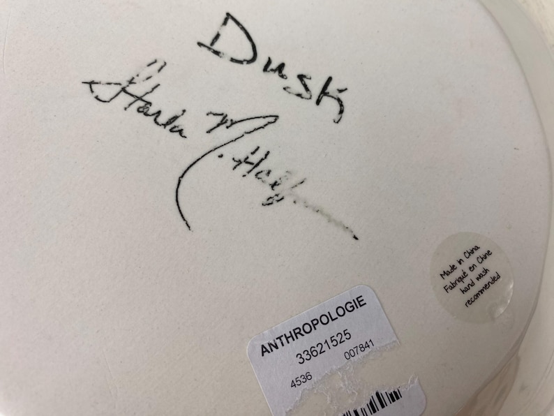 May include: Close-up of a white ceramic plate with black handwritten text that reads "Dusk" and a signature. A sticker with the word "ANTHROPOLOGIE" and other details is visible.