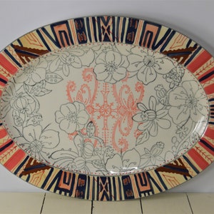 May include: Oval ceramic serving platter with a white background and a floral design in black. The border is decorated with a geometric pattern in red, orange, blue, and brown.