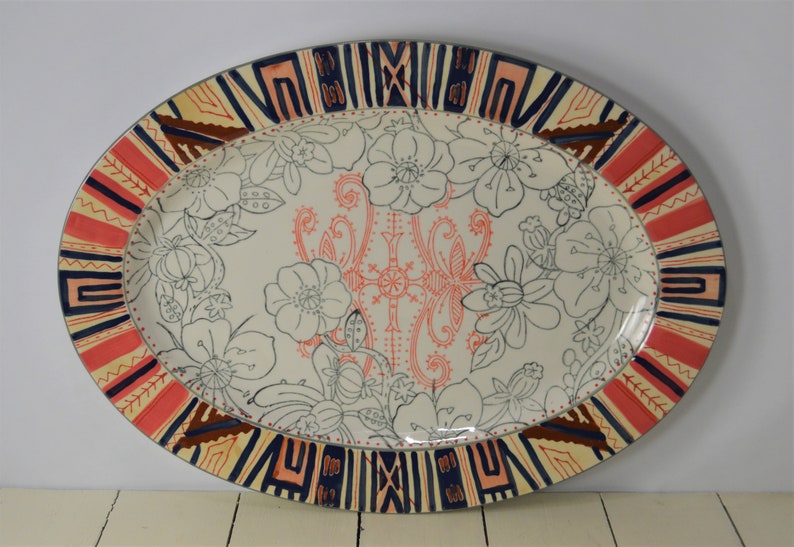 May include: Oval ceramic serving platter with a white background and a floral design in black. The border is decorated with a geometric pattern in red, orange, blue, and brown.