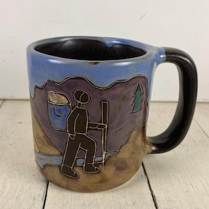 May include: A ceramic mug featuring a hand-drawn illustration of a hiker with a backpack and walking stick. The design includes a purple mountain range, a green pine tree, and a light blue sky. The mug has a dark brown handle and interior.