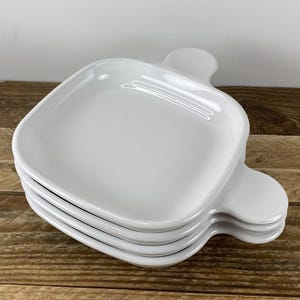 May include: A stack of four white ceramic serving dishes. Each dish has a square shape with rounded corners and a handle on one side. The dishes are stacked on a wooden surface.