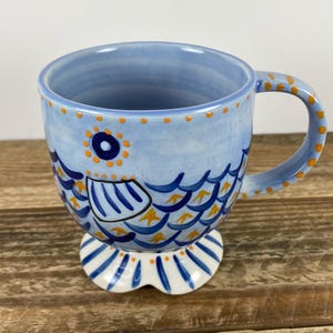 Op de afbeelding: Een keramische mok met een grillig visontwerp. De mok is voornamelijk lichtblauw met oranje en donkerblauwe accenten. De visillustratie loopt rond de mok en de handgreep en voet zijn ook versierd.