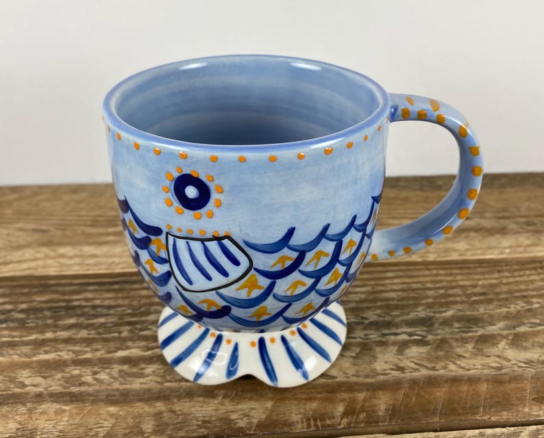 Op de afbeelding: Een keramische mok met een grillig visontwerp. De mok is voornamelijk lichtblauw met oranje en donkerblauwe accenten. De visillustratie loopt rond de mok en de handgreep en voet zijn ook versierd.
