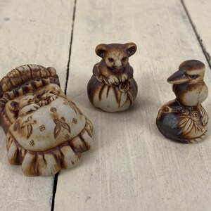May include: Three ceramic figurines: a sleeping baby in a bed, a koala bear, and a bird. The figurines are primarily brown and cream-colored, with detailed carvings. The koala and bird are perched on decorative bases.