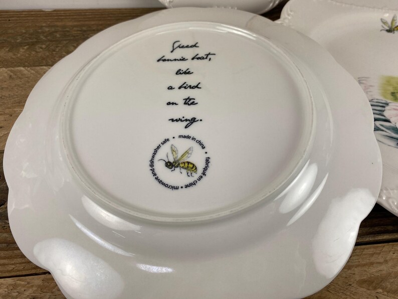 May include: White ceramic plate with a scalloped edge. The plate features a black inscription that reads "Freed Lonnie beat, like a bird on the wing." A yellow and black bee illustration is also present, along with text indicating it is microwave and dishwasher safe.