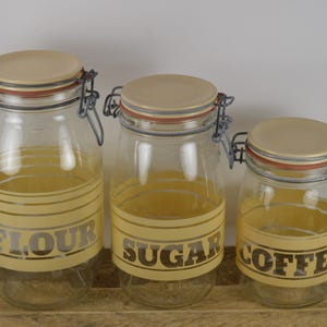 May include: Three glass canisters with beige lids and yellow striped labels. The labels read "Flour", "Sugar", and "Coffee".