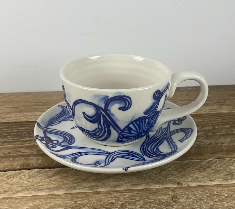 Puede incluir: Juego de taza y platillo de t&eacute; de cer&aacute;mica blanca con dise&ntilde;os florales abstractos azules. La taza de t&eacute; tiene un asa y el platillo es redondo. Los dise&ntilde;os azules est&aacute;n pintados en el exterior de la taza y el platillo.