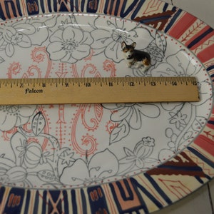 May include: A white oval platter with a floral design in black and red. The platter has a decorative border in shades of red, blue, and cream. A small figurine of a black and brown dog is sitting on the platter.