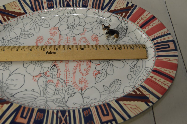 May include: A white oval platter with a floral design in black and red. The platter has a decorative border in shades of red, blue, and cream. A small figurine of a black and brown dog is sitting on the platter.