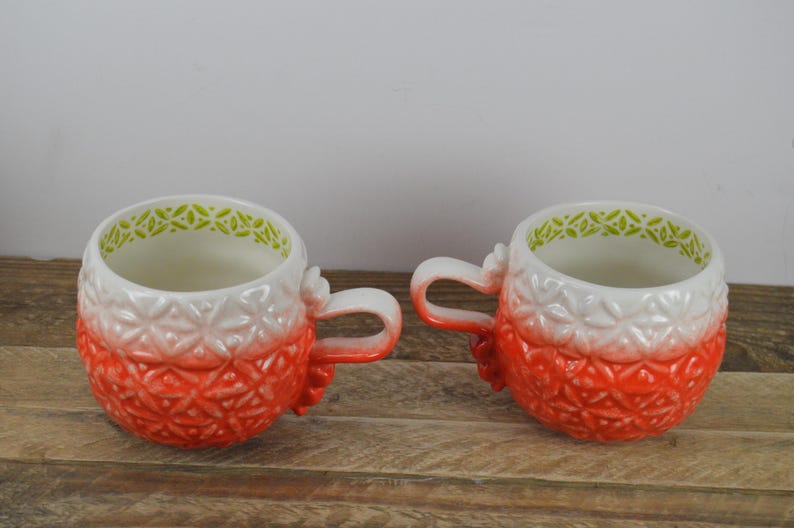 May include: Two red and white ceramic mugs with green trim and a textured design. The mugs have a gradient effect from white to red.