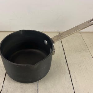 May include: A small, black saucepan with a long, silver handle. The pan has a rounded body and a pouring spout. The handle is attached with two rivets. The pan is sitting on a white wooden surface.