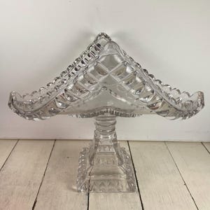May include: A clear, vintage glass cake stand with a diamond pattern on the top and a scalloped edge. The stand has a square base and a pedestal stem. The cake stand is displayed on a white wooden surface.
