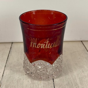 May include: A vintage red glass with a clear, cut-glass base. The word "Monticello" is written in gold script on the side of the glass. The glass has a gold rim.