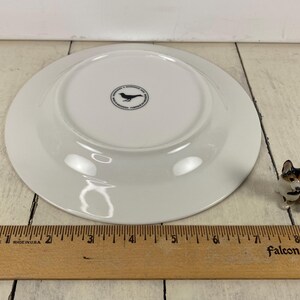 May include: A white ceramic plate with a black bird logo on the underside. A small corgi figurine is to the right of the plate. A wooden ruler with inch measurements is in the foreground.