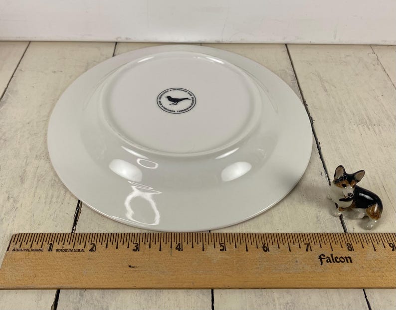 May include: A white ceramic plate with a black bird logo on the underside. A small corgi figurine is to the right of the plate. A wooden ruler with inch measurements is in the foreground.
