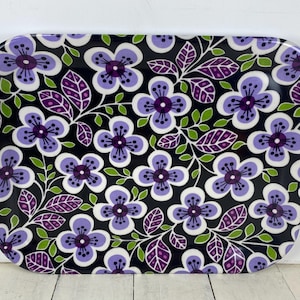 May include: A rectangular serving tray with rounded corners featuring a repeating floral pattern. The design consists of light purple flowers with dark purple centers, interspersed with dark purple and green leaves against a solid black background.
