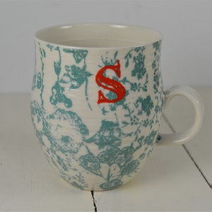 May include: White ceramic mug with a blue and white floral pattern and a red letter "S" on the front.