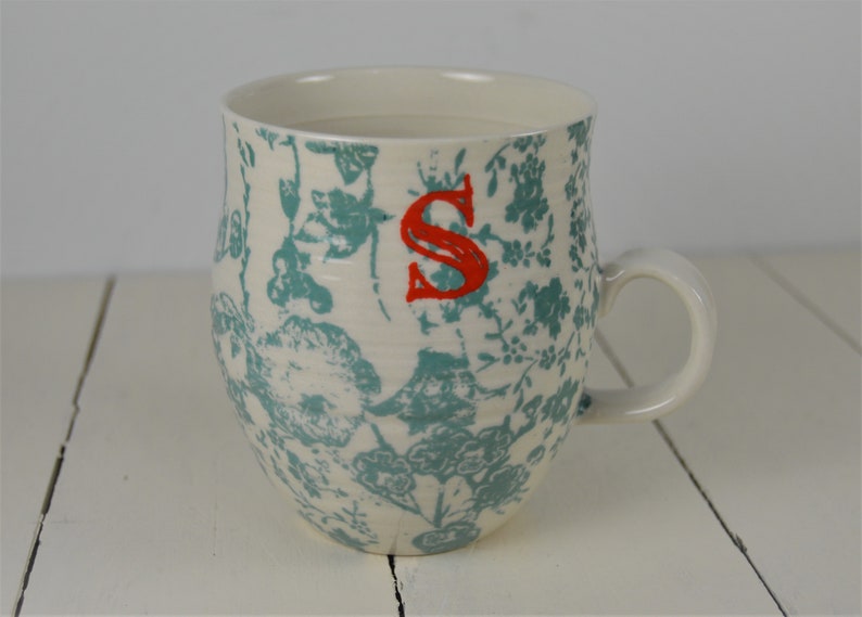 May include: White ceramic mug with a blue and white floral pattern and a red letter "S" on the front.