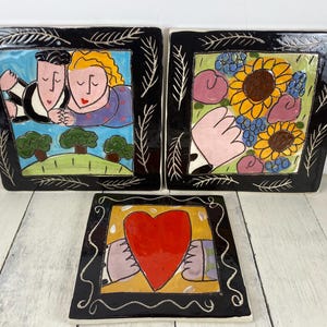 May include: Three square ceramic art pieces with black frames. One features a couple, another sunflowers, and the third a heart. Each has a unique, colorful, hand-painted design. The frames have a white leaf-like pattern.