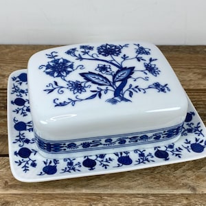 May include: White ceramic butter dish with a blue floral design. The lid features a detailed tree and flower pattern, while the base has a repeating floral and fruit motif. The dish is square with rounded corners, and the design is reminiscent of classic blue and white china.