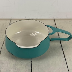 May include: A teal enamelware saucepan with a white interior and a black rim. The saucepan has a single handle and a pouring spout. This vintage kitchenware is perfect for cooking and serving.